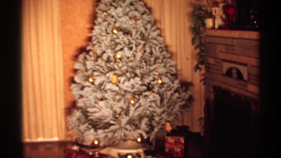Wisconsin 1971 home-movie frame showing a flocked tree with small lights, drapery, a fireplace, and holiday packages in a private living room.