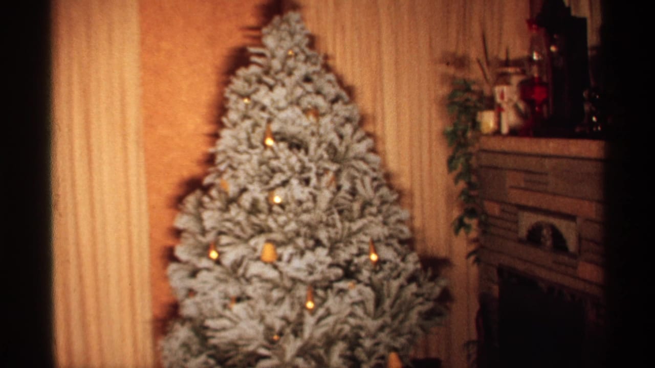 1971 Wisconsin home-movie frame showing a snow-flocked Christmas tree with gold lights near a fireplace mantel and paneled wall.