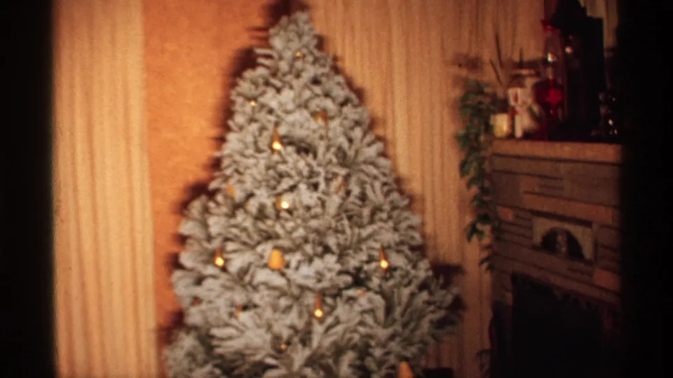 1971 Wisconsin home-movie frame showing a snow-flocked Christmas tree with gold lights near a fireplace mantel and paneled wall.
