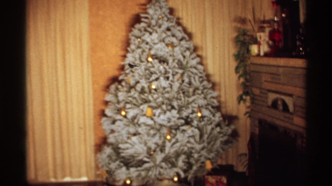 1971 Wisconsin home-movie frame with the flocked artificial Christmas tree centered near a curtained wall, with a fireplace mantel and holiday objects visible to the right.