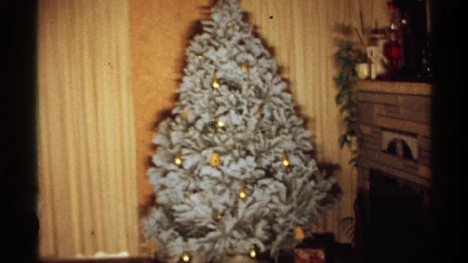 1971 Wisconsin 8mm frame showing a closer, slightly soft view of the white-flocked Christmas tree, yellow ornaments, glowing lights, and the fireplace mantel in a private home.