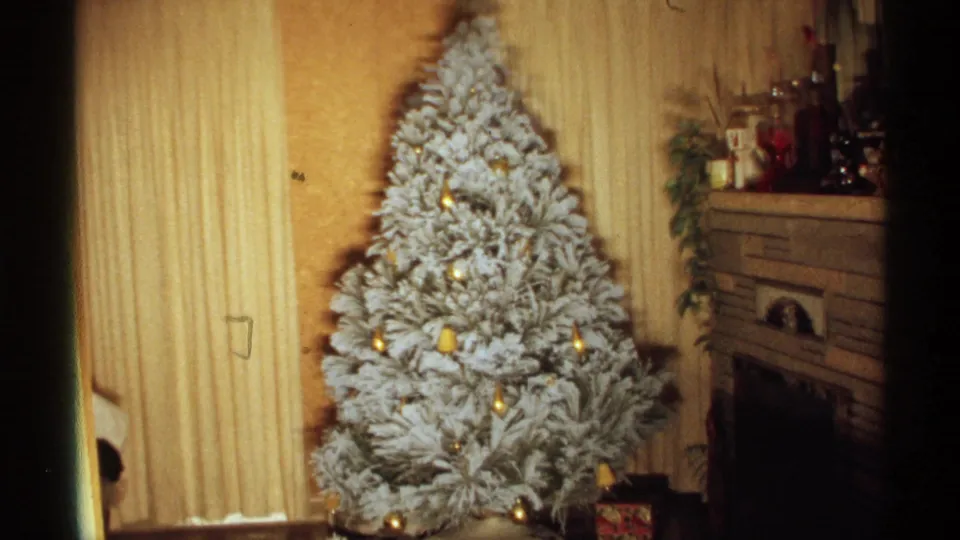 1971 Wisconsin home-movie frame showing the large flocked tree with lights and ornaments, a curtained window or wall area, and the fireplace mantel at the right edge.