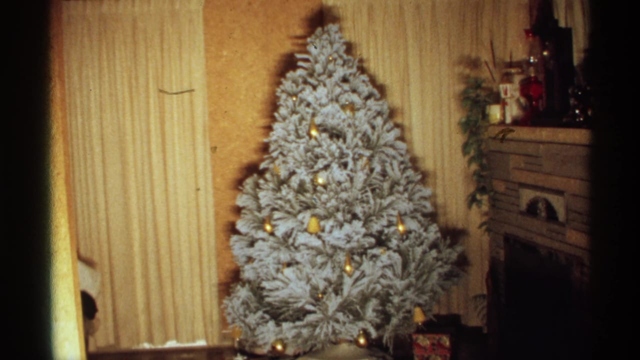 White frosted Christmas tree with gold ornaments beside vertical blinds and a fireplace.