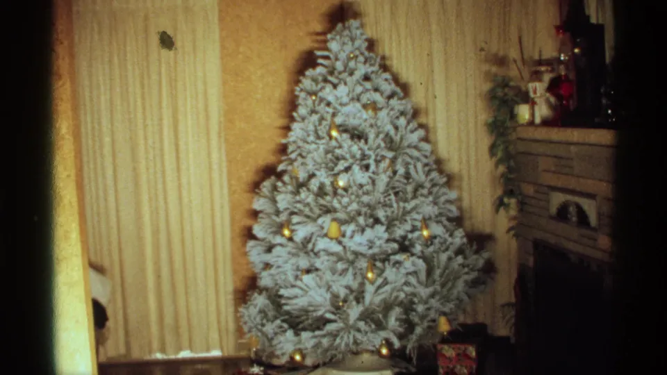 1971 home-movie frame of a flocked Christmas tree in a Wisconsin living room with gold ornaments, blinds, and mantel decor.