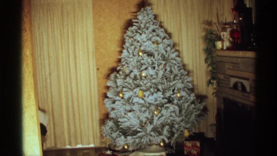 White frosted Christmas tree with gold ornaments remains static in a 1971 Wisconsin living room near a fireplace and blinds.
