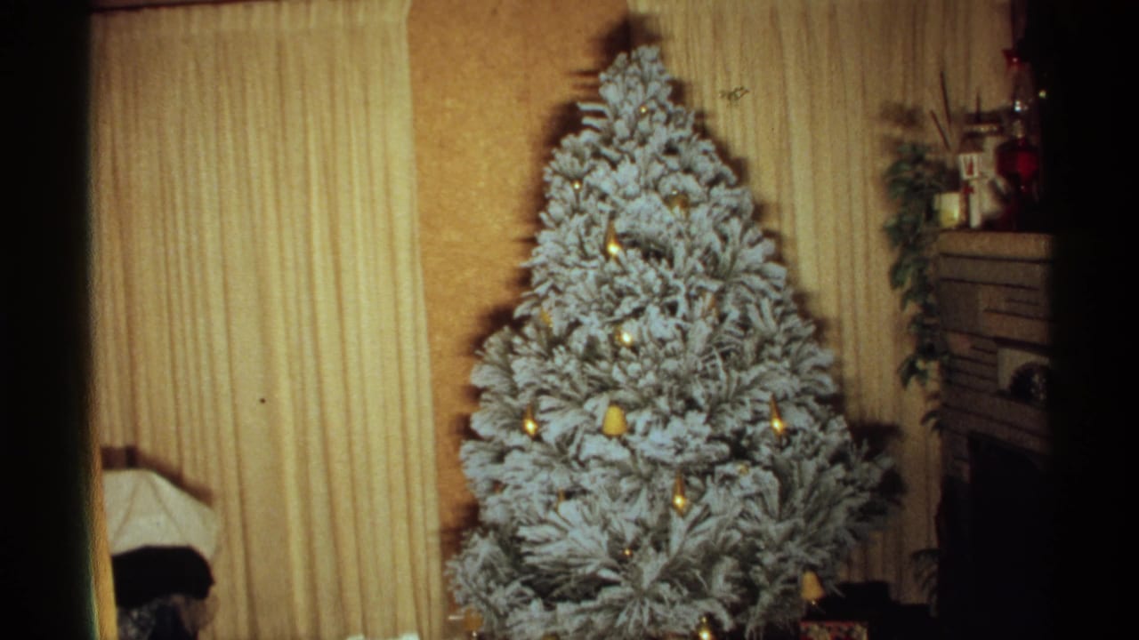 Flocked Christmas tree indoors beside yellow curtains and a fireplace mantel.