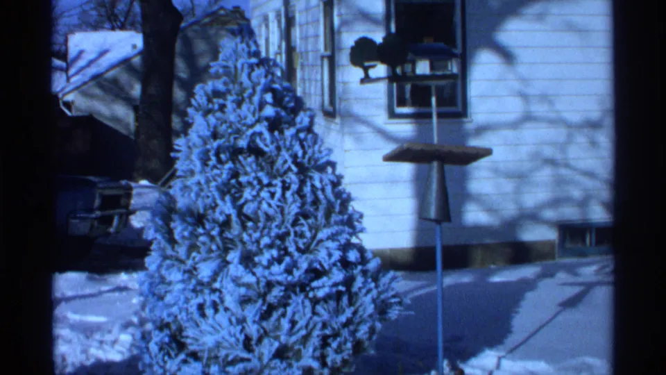 1971 Wisconsin 8mm frame showing the flocked tree outdoors in a snowy yard beside a house wall, window, and bird feeder pole.