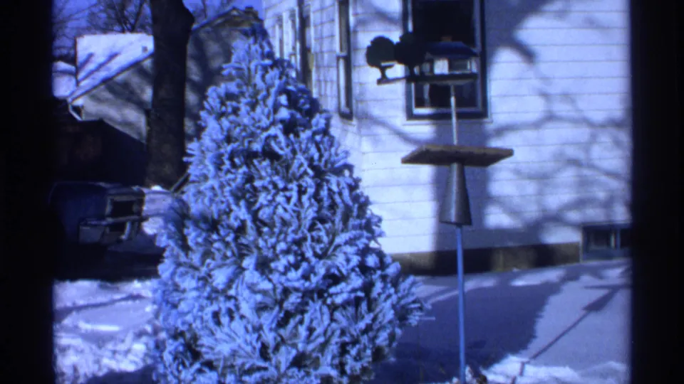 1971 Wisconsin 8mm frame showing a small evergreen heavy with snow outside a white house, with a bird feeder and partial car visible.