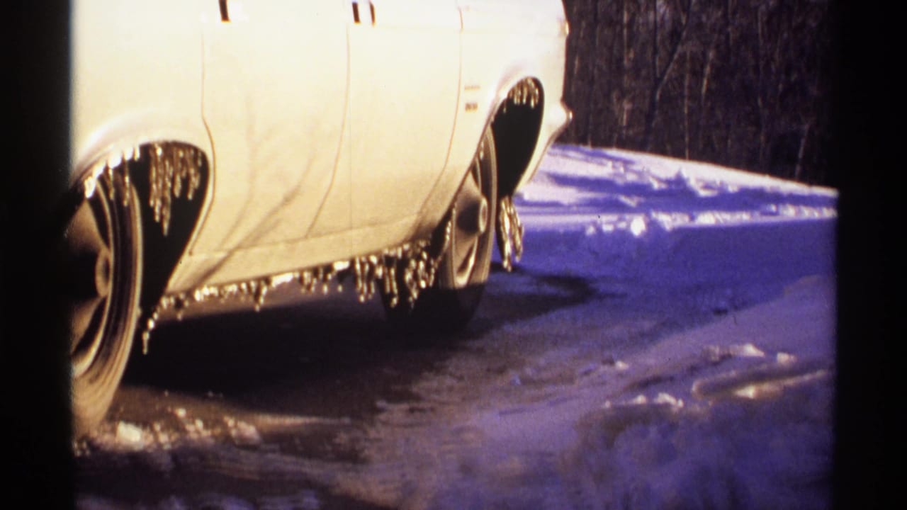 White car parked beside snow with icicles hanging from the fender and wheel well.