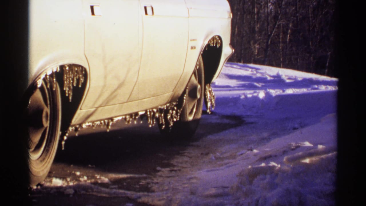 Side of a white car with icicles along the lower body beside blue-toned snow.