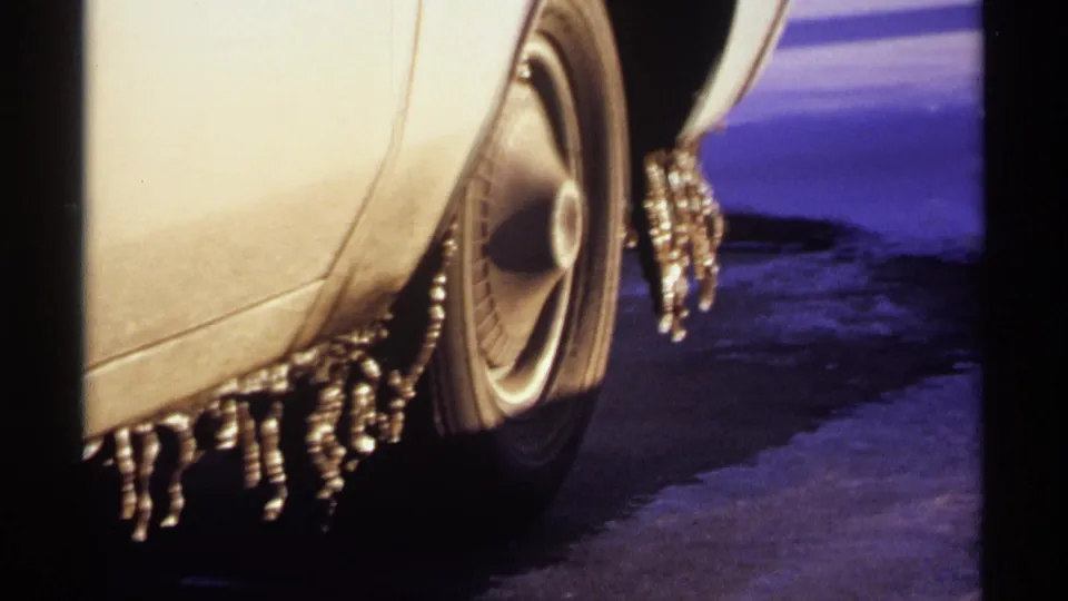 Closer 1971 Wisconsin 8mm view of a white car's wheel and lower fender, with icicles hanging over snow and ice.