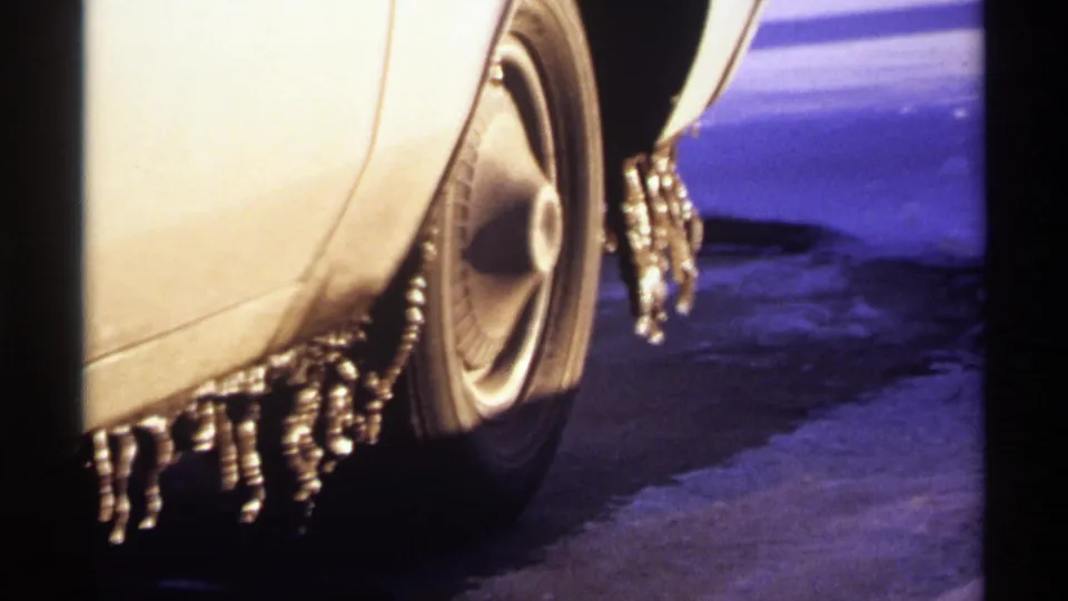 1971 Wisconsin 8mm frame of the car wheel and fender, with icicles hanging against blue-toned snow and dark roadside shadows.