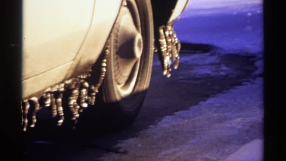 1971 Wisconsin home-movie frame showing a close-up of an older car wheel, fender, and hanging icicles on pavement.