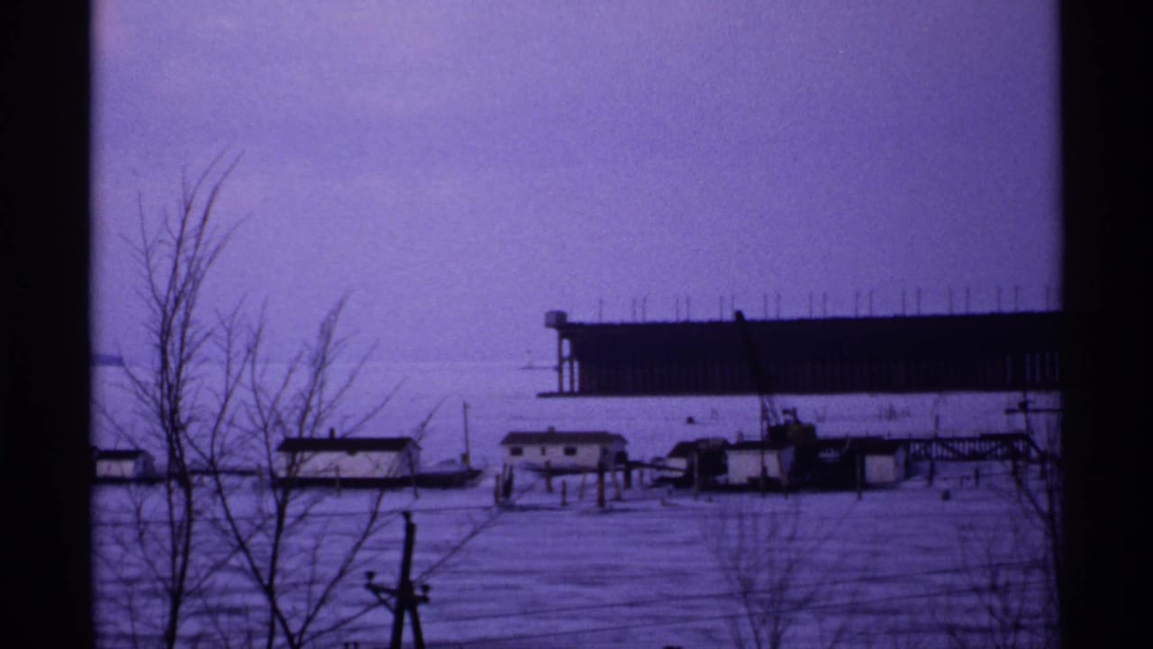 1971 Wisconsin 8mm frame of a snow-covered waterfront with bare trees, small shoreline buildings, and a large dark structure beyond the ice or water.