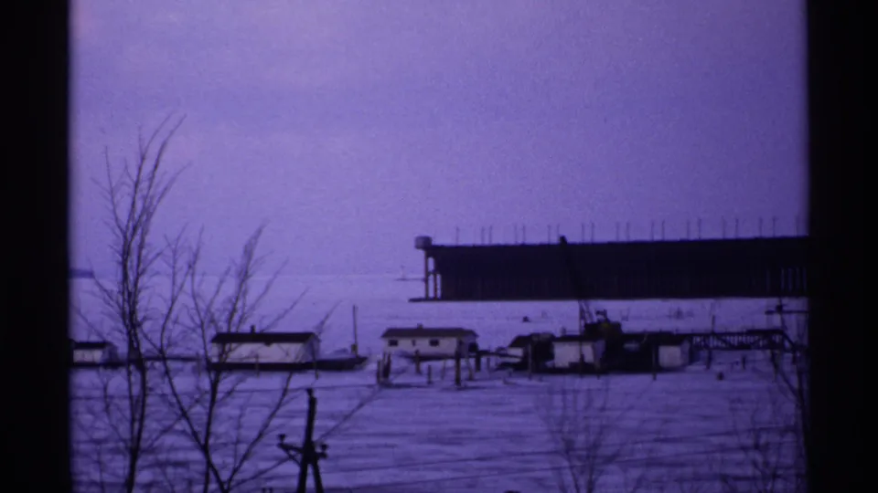 1971 Wisconsin 8mm frame of a snow-covered waterfront with bare trees, small shoreline buildings, and a large dark structure beyond the ice or water.