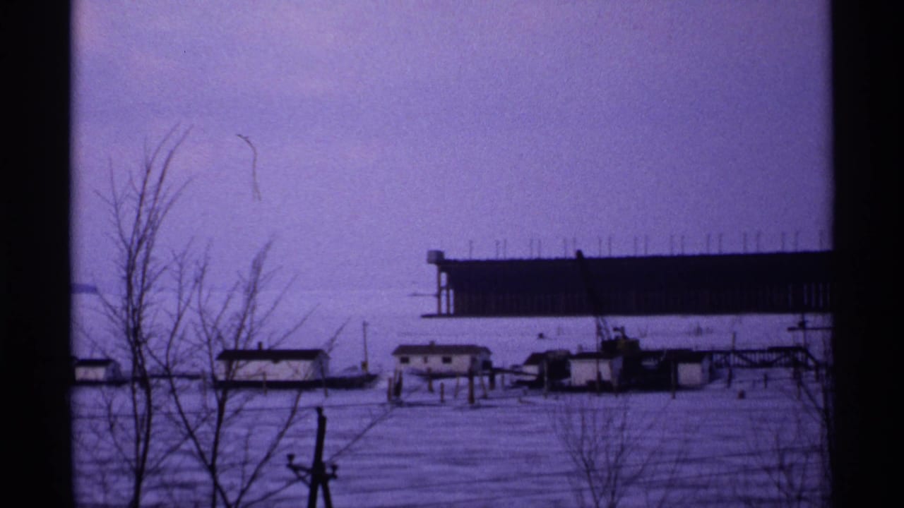 1971 Wisconsin winter frame showing a frozen or snow-covered waterfront, leafless trees, low buildings, and a long dark pier-like structure in the background.