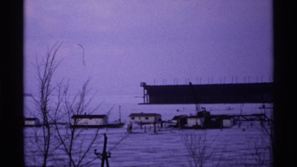 1971 Wisconsin winter frame showing a frozen or snow-covered waterfront, leafless trees, low buildings, and a long dark pier-like structure in the background.