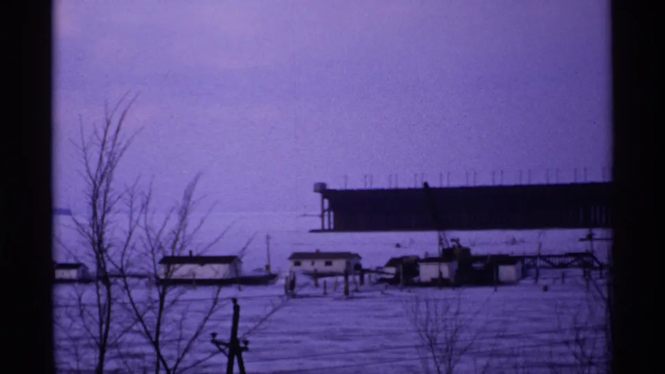Soft 8mm winter view in Wisconsin, 1971, with snow-covered ground, bare foreground trees, small waterfront structures, and a distant dark industrial form.