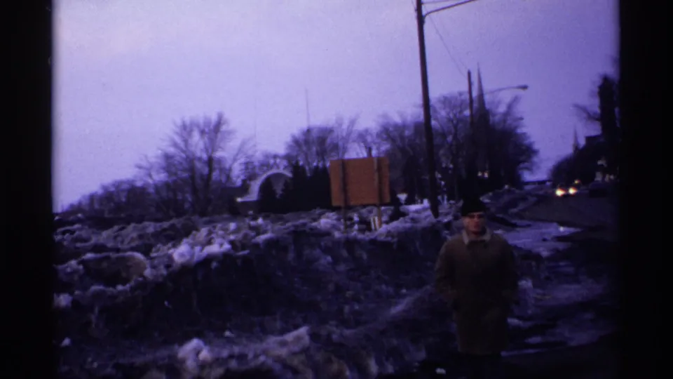1971 Wisconsin street-side frame with an adult man in a long winter coat near snowbanks, roadside signs, utility poles, bare trees, and passing car lights.