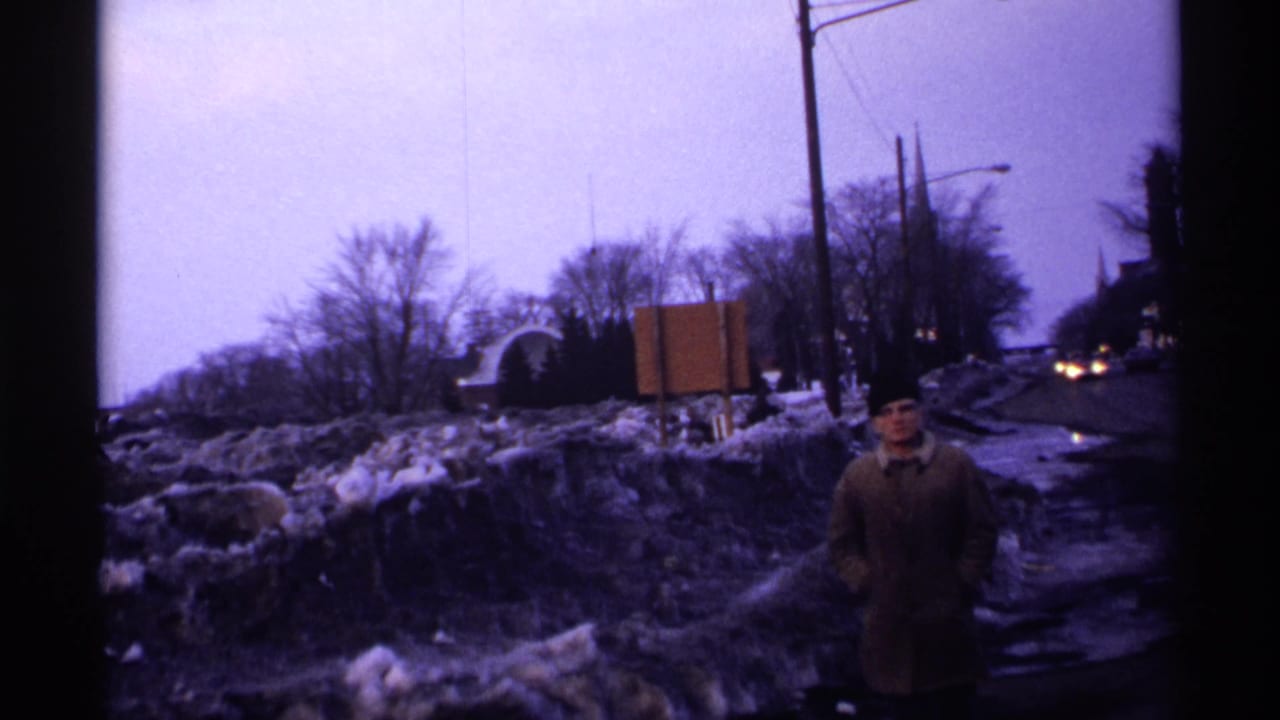 Man in a winter coat stands near a snowy roadside with bare trees and road signs behind him.