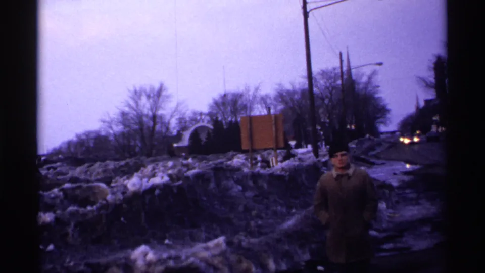 1971 Wisconsin 8mm frame showing a man in a heavy coat and hat by slushy roadside snowbanks, with bare trees, signs, and wet pavement nearby.