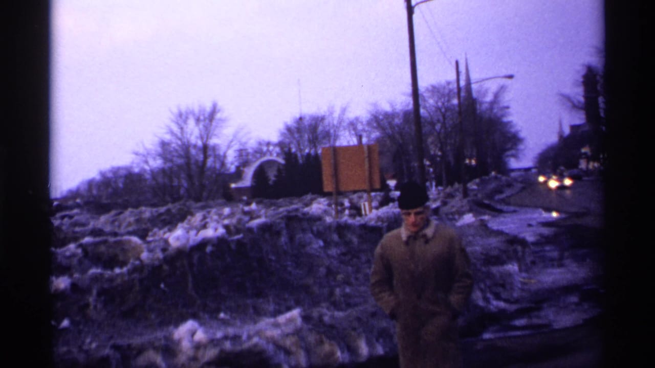 1971 Wisconsin roadside view with a bundled man near dirty snowbanks, wet pavement, bare winter trees, and distant car headlights.