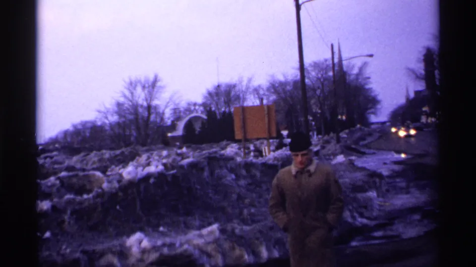 1971 Wisconsin roadside view with a bundled man near dirty snowbanks, wet pavement, bare winter trees, and distant car headlights.