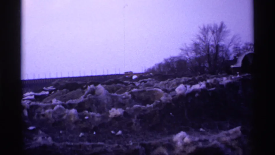 1971 Wisconsin 8mm winter roadside landscape after the man leaves the frame, showing slushy snowbanks, bare trees, road edge, and distant traffic.