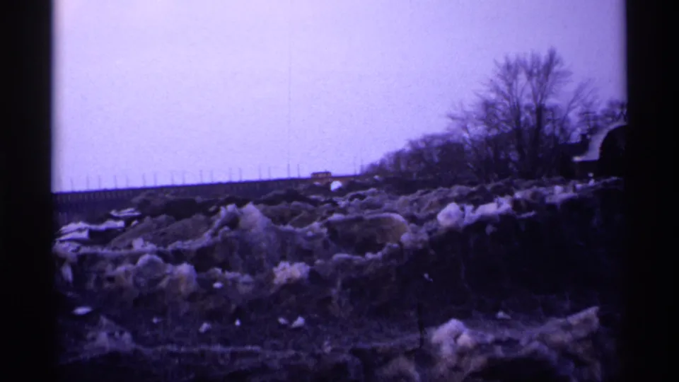 1971 Wisconsin 8mm frame showing a quiet winter roadside with dirty snowbanks, bare trees, a distant road, and muted overcast light.