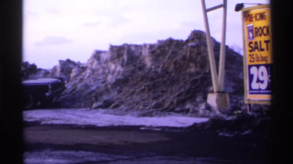 1971 Wisconsin winter frame with a yellow rock salt sign advertising 29 cents, a dirty snow pile, and a parked dark car.