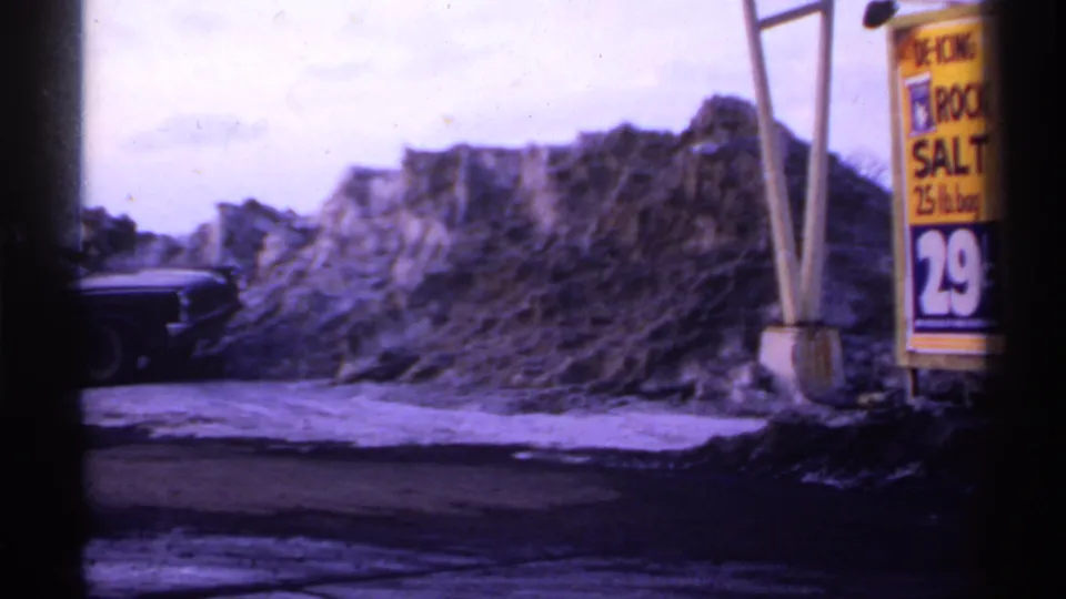 1971 Wisconsin 8mm view of a roadside snow pile with a partial dark car, wet pavement, poles, and a yellow rock salt sign.