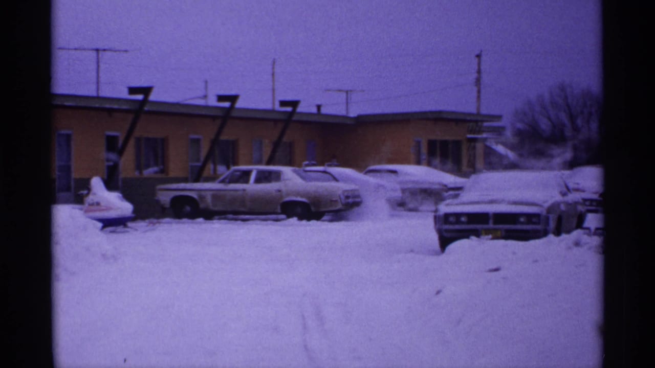 Snowy motel lot with several parked cars and a front-facing car at right.