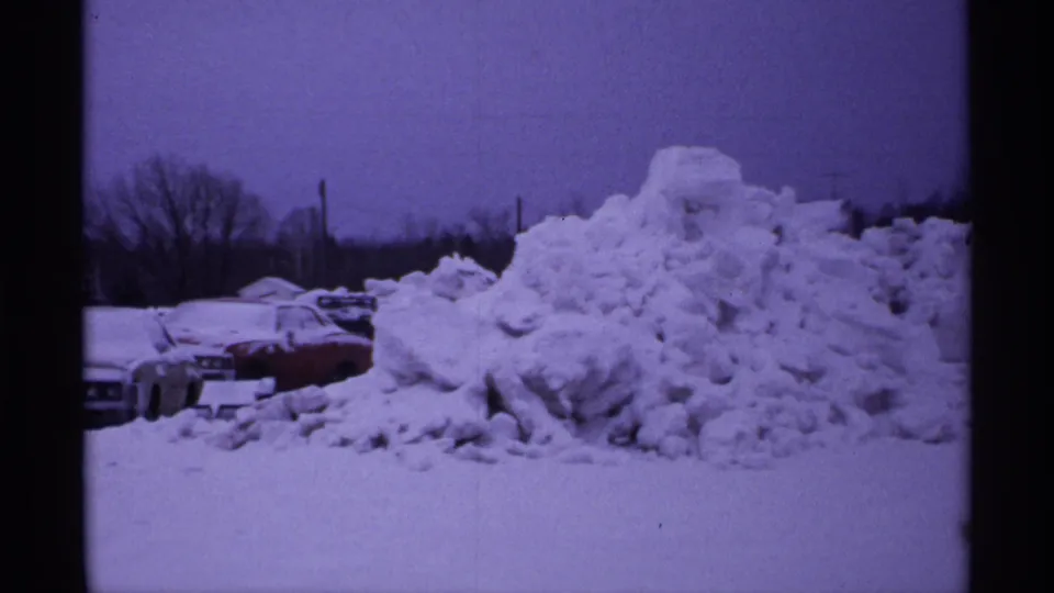 1971 Wisconsin frame showing a high mound of plowed snow beside partially visible cars in a winter motel parking lot.