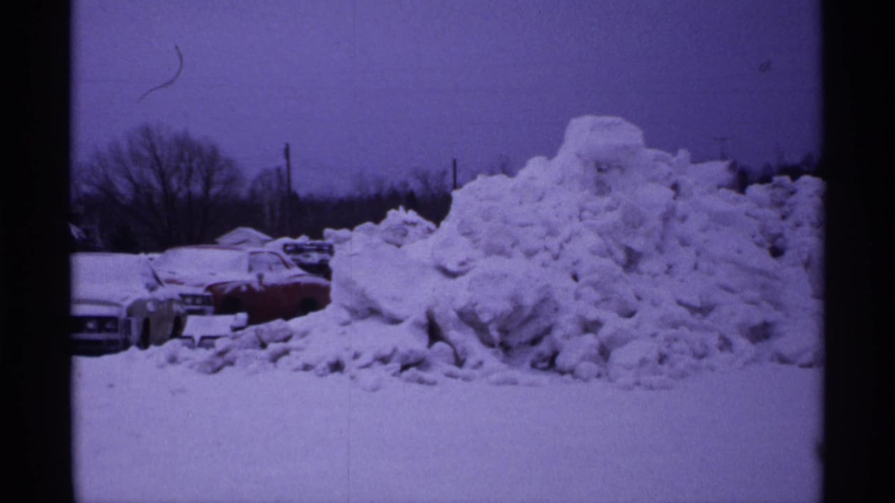 Snow-covered parked cars beside a large plowed snow pile under a gray sky.