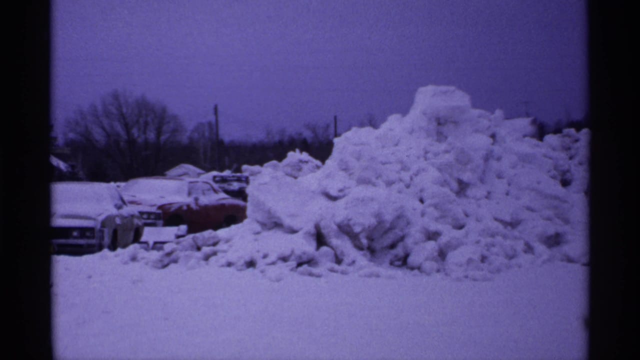 Large snowbank with partially visible parked cars and bare trees behind it.