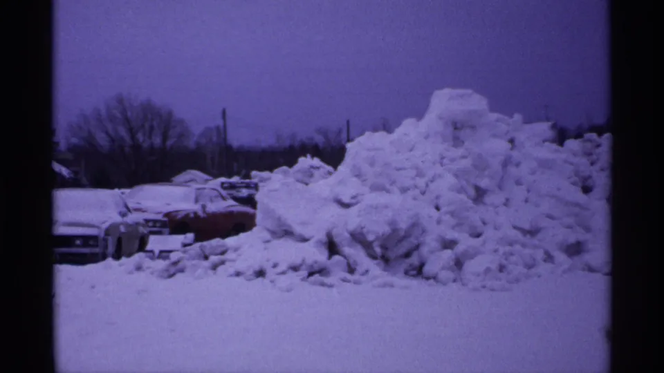 1971 Wisconsin winter frame with a high snow pile dominating the foreground and parked cars partly visible at the left.