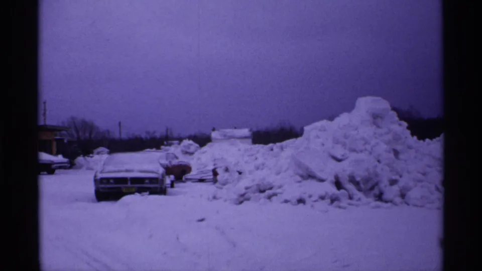 1971 Wisconsin home-movie frame showing a parked car front-on beside a plowed snow pile, with winter trees and utility poles in back.