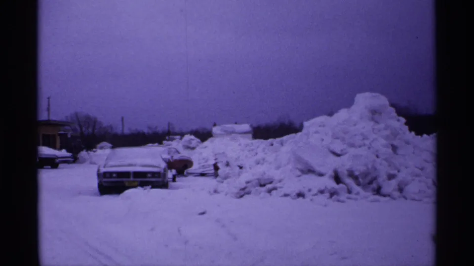 1971 Wisconsin winter frame showing a parked car, another vehicle, and a broad snowbank across a snowy lot.