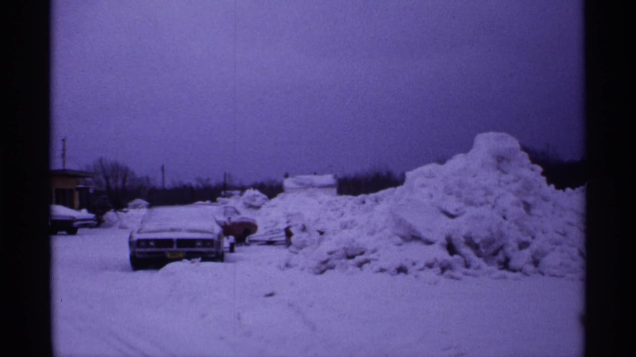 Snow-covered parked cars beside a large plowed snowbank at dusk.