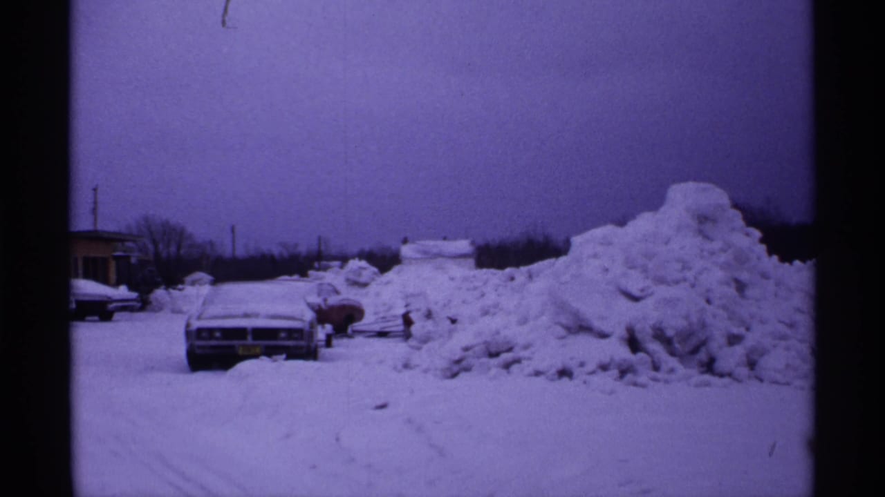 Parked sedan and snowbank seen across a snowy lot or street.