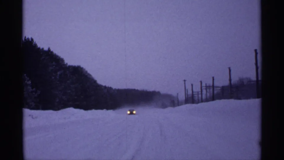 1971 Wisconsin 8mm frame showing a car with headlights on traveling along a snow-covered road at dusk.