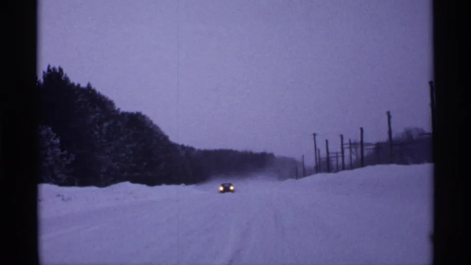 1971 Wisconsin winter frame with a distant car driving through a snowy road corridor under a dim sky.