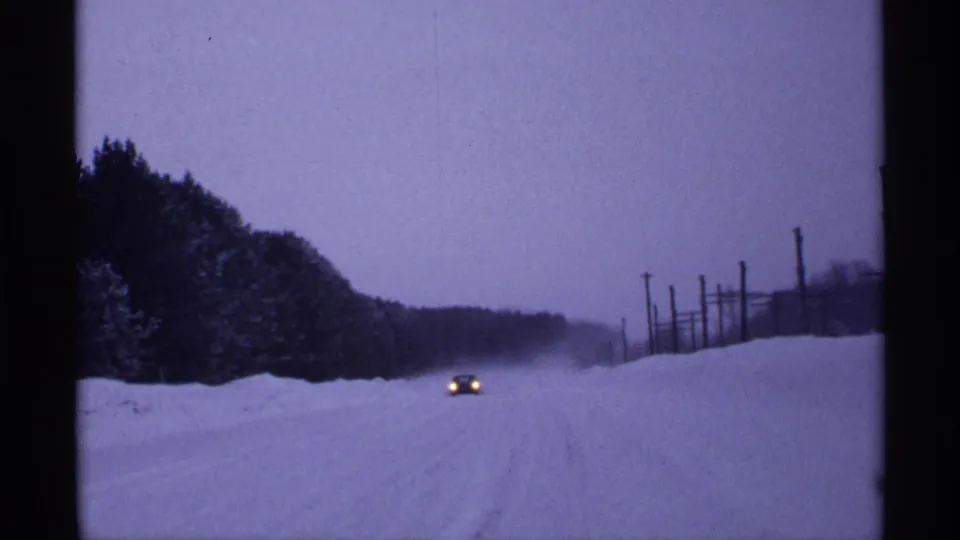 In 1971 Wisconsin 8mm footage, a distant car approaches along a snowy rural road lined with trees and utility poles.