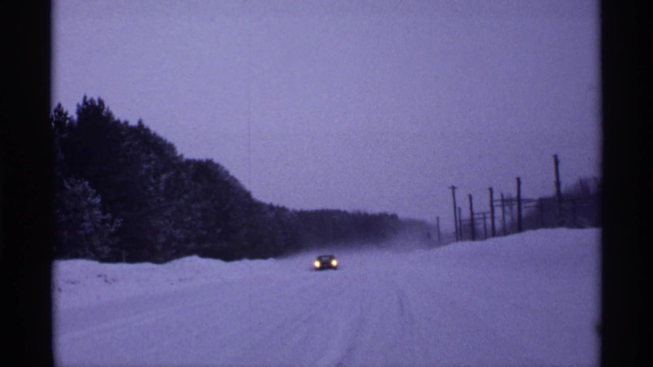 Car with headlights visible on a snowy road bordered by trees and poles.