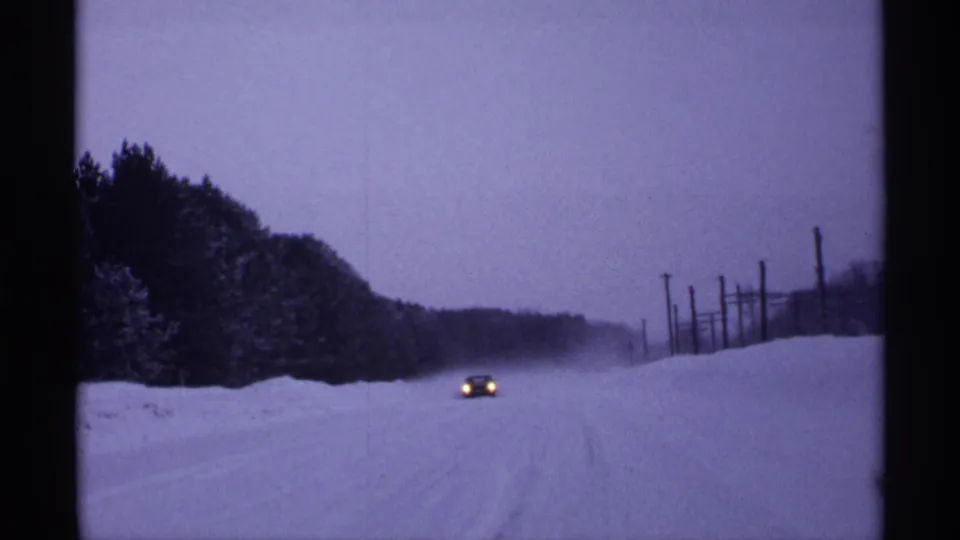 A small car continues along a snow-covered Wisconsin rural road, its headlights showing through dim winter light and snowfall.