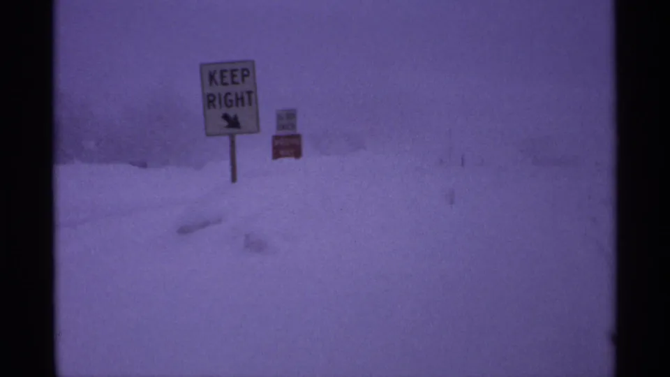Snowy 1971 home-movie frame showing public road signs against a pale winter sky near Ironwood.