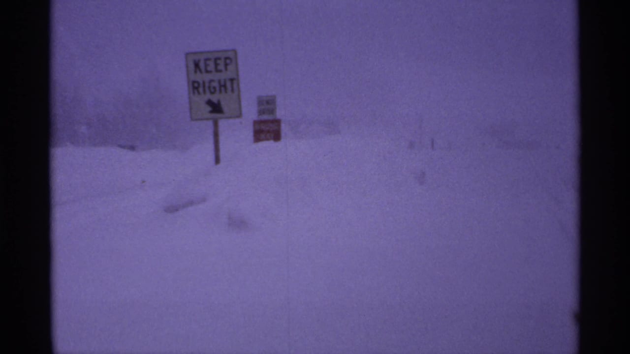 Snow-covered roadside with a KEEP RIGHT sign and smaller red sign in low visibility.
