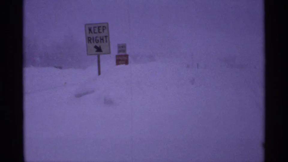 1971 Wisconsin 8mm frame showing deep snow, a KEEP RIGHT sign, and a smaller red roadside sign during a blizzard.