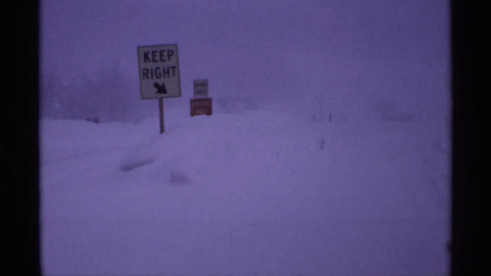 Vintage 8mm view of Wisconsin roadside signs partly buried by snow during a heavy winter storm.