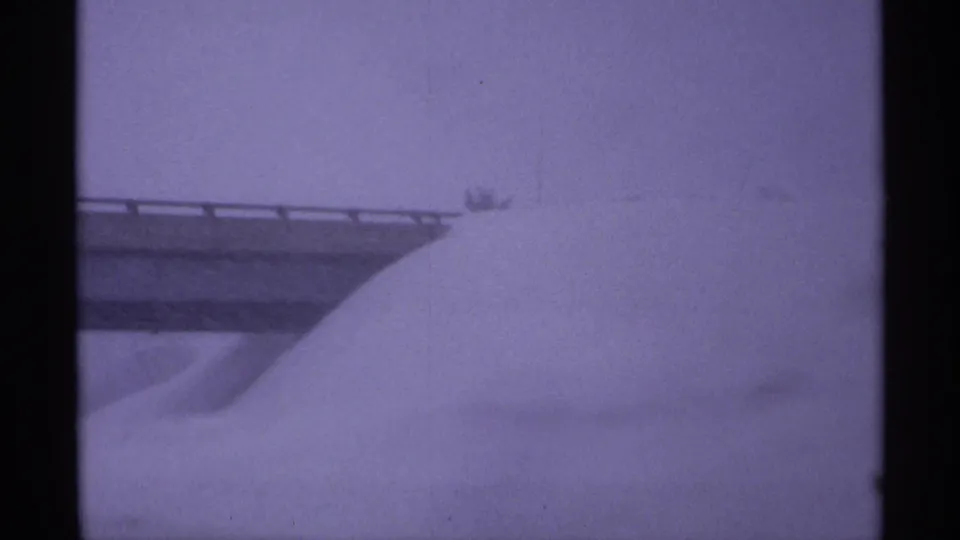 Closer 8mm view of an overpass guardrail and a large snowdrift rising below it in Wisconsin winter conditions.